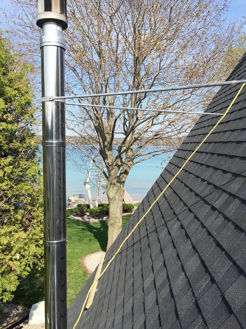 Chimney on a roof with shingles, lake and trees in the background. A yellow rope is on the roof.