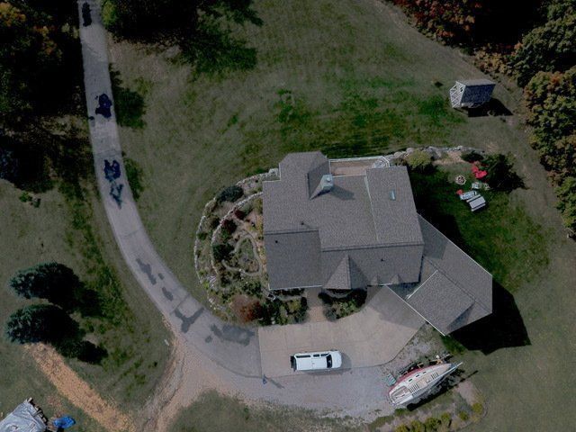 Aerial view of a house with a curved driveway and vehicles, surrounded by grass and trees.