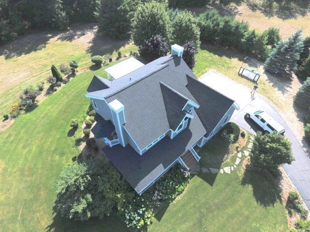 Aerial view of a gray house with a dark roof on a grassy hill; a car is parked nearby.