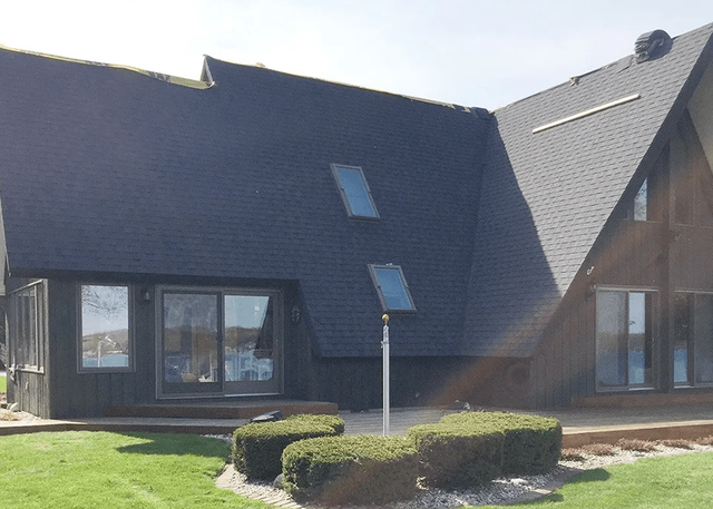 Dark, angled roof on a waterfront home with sliding glass doors, skylights, and manicured bushes on the lawn.