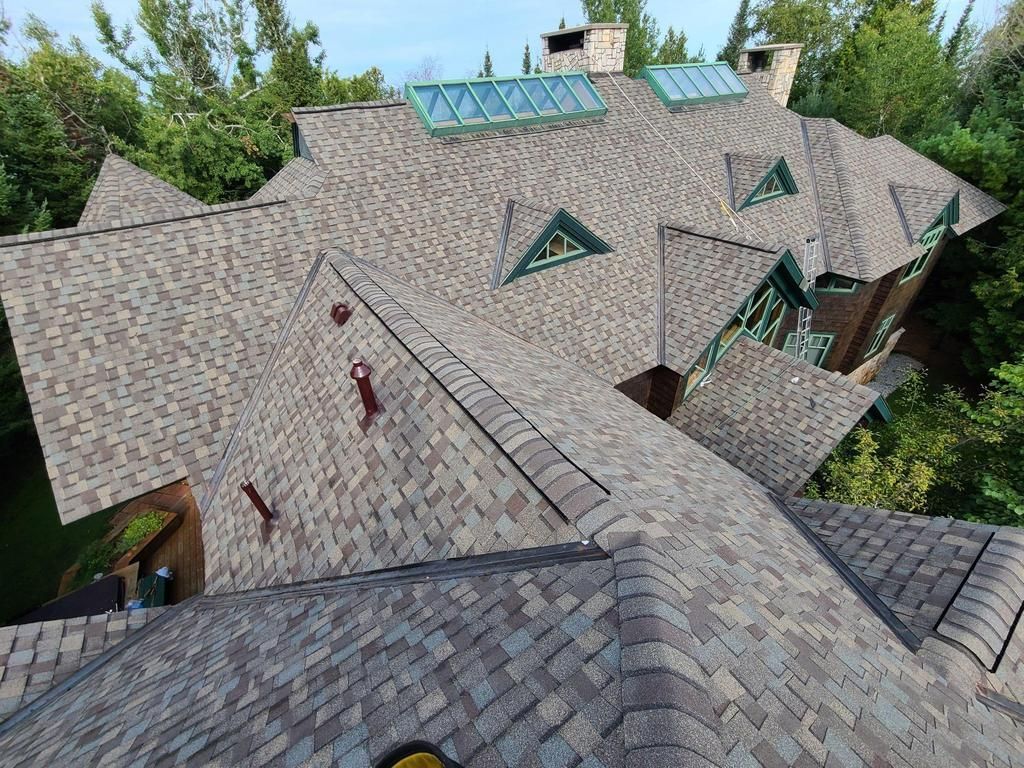 Brown asphalt shingle roof on a large house with multiple gables, skylights, and green trim, surrounded by trees.