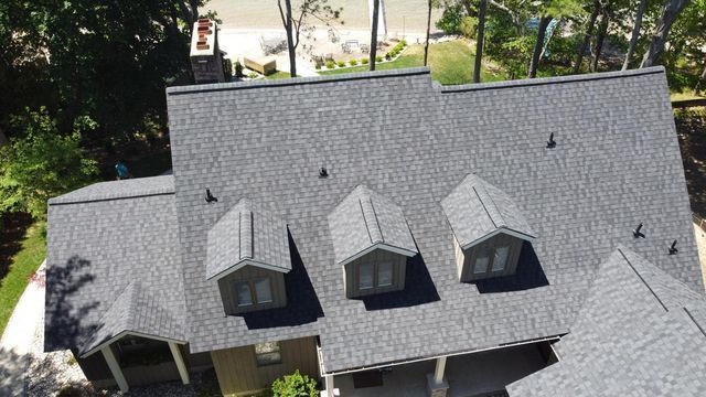 Gray shingle roof of a house with three dormers, trees and water in the background.