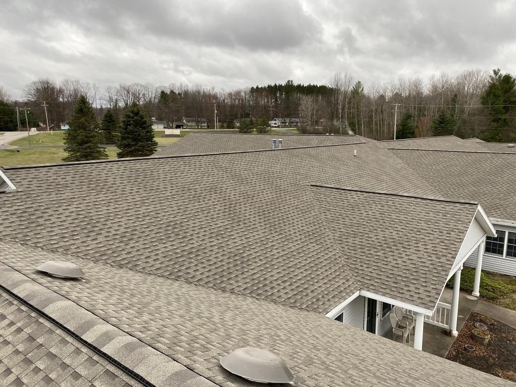 Gray asphalt shingle roofs of multiple buildings, overcast sky, trees in the distance.