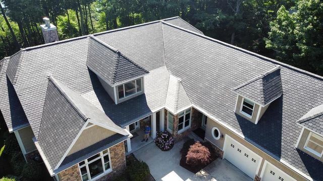 Aerial view of a house with a complex gray roof, surrounded by trees. Beige walls and a garage.