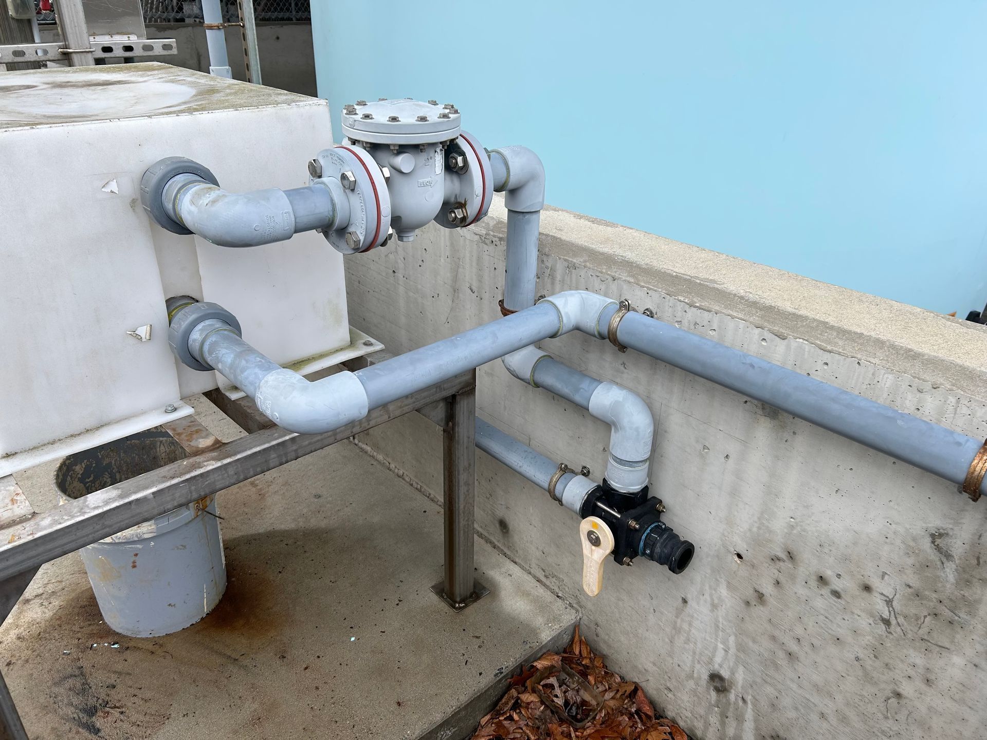 Industrial plumbing pipes and valves along a concrete wall, with a rust-stained bucket below