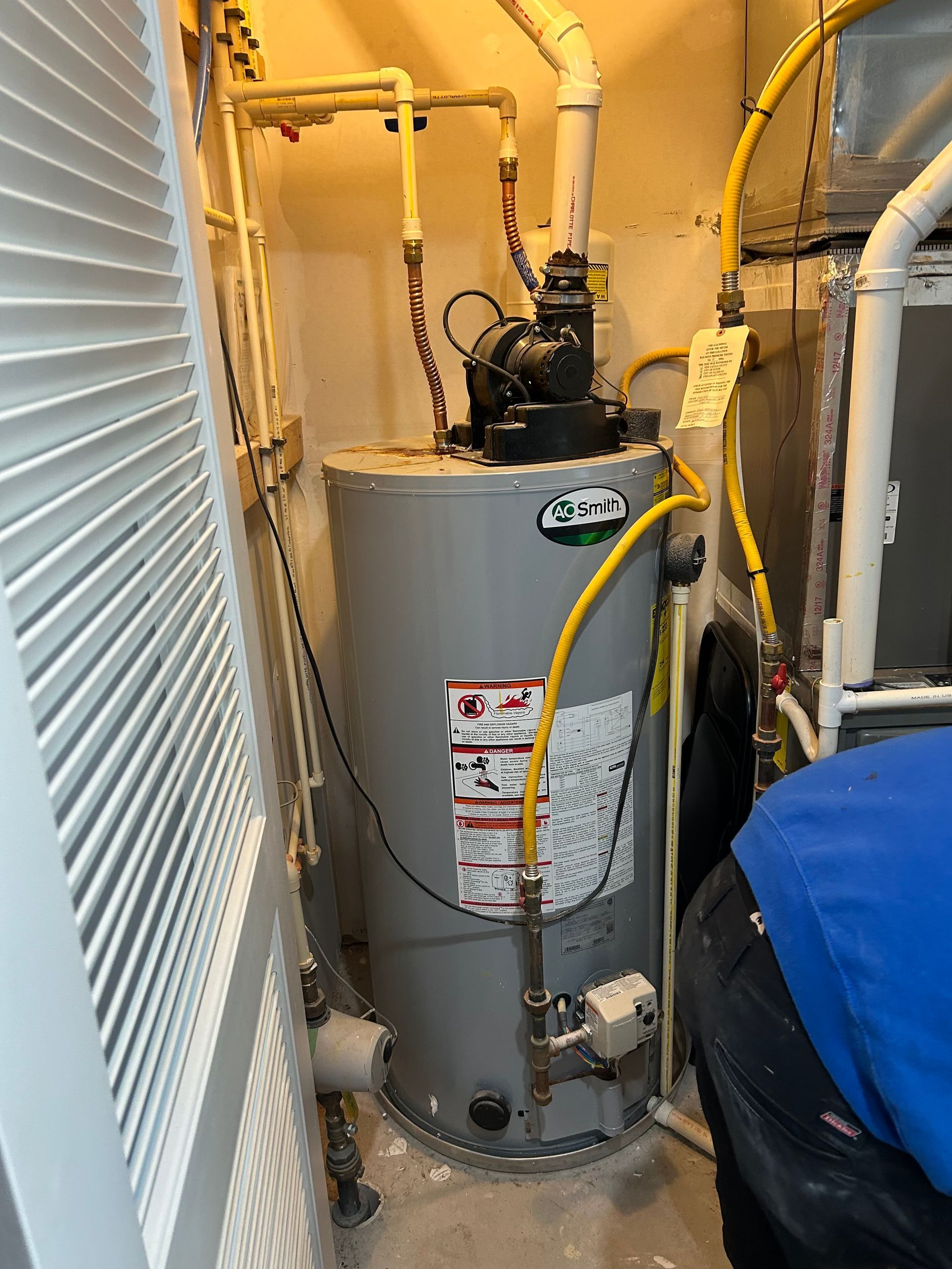 Water heater in a narrow utility closet, with pipes and hoses attached; a person in blue is partly visible on the right