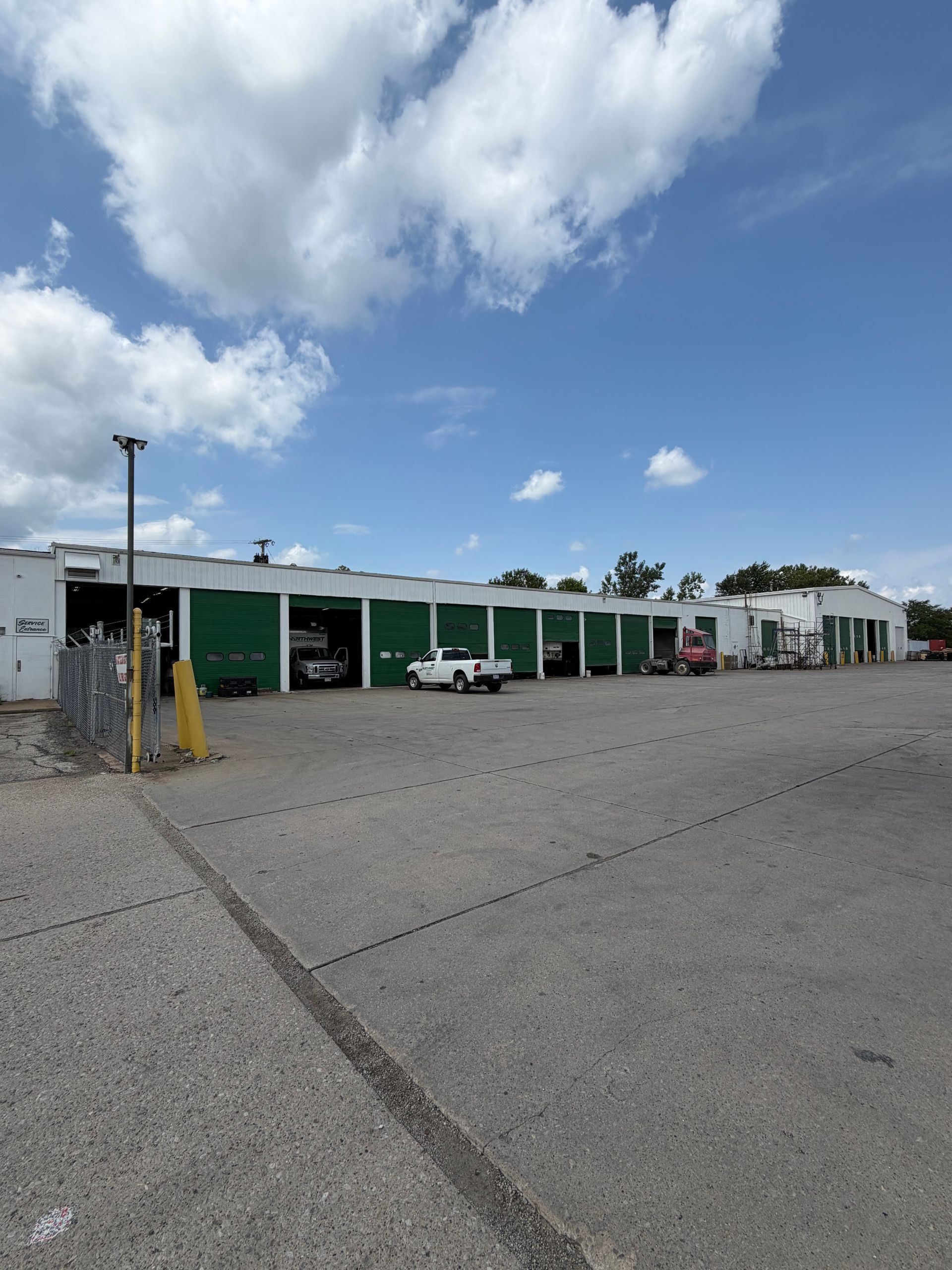 Low industrial building with green awnings, parked vehicles, and a cloudy sky over a gravel lot
