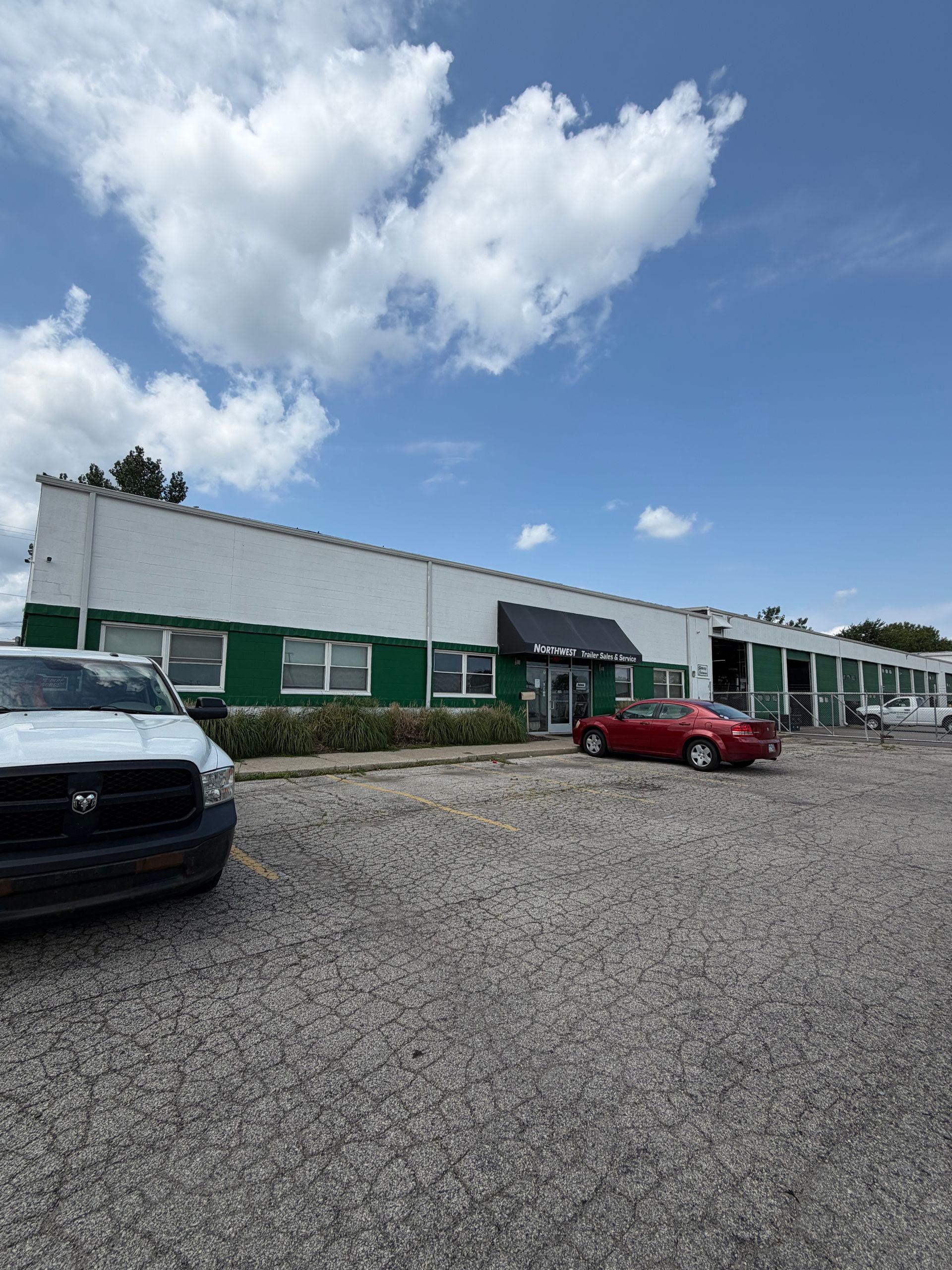 Industrial building with green-trim windows, gravel lot, and parked red and black cars under a partly cloudy sky