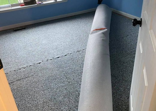 Gray fabric roll leaning against a blue wall on a carpeted floor in a room.