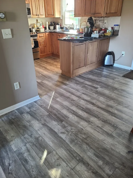 Empty kitchen with wood cabinets and a large island, viewed from a room with wood-look flooring.
