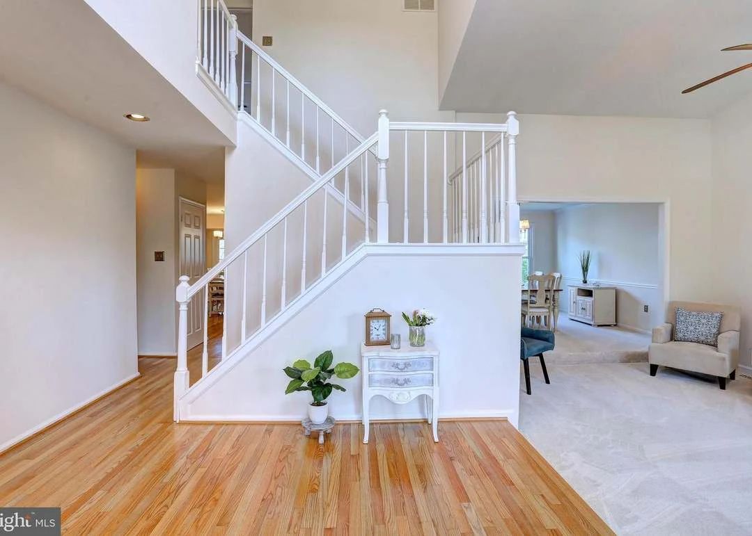 Bright foyer with wood floors, white staircase, and open living room with beige seating