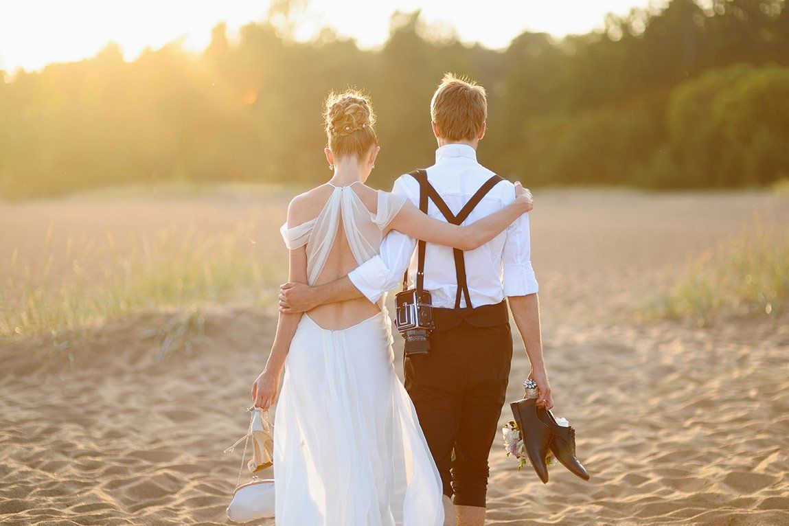  A newlywed couple - a man and a woman - walk across a beach at sunset.