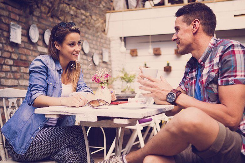 A heterosexual couple smiles at each other while seated at a café table. 