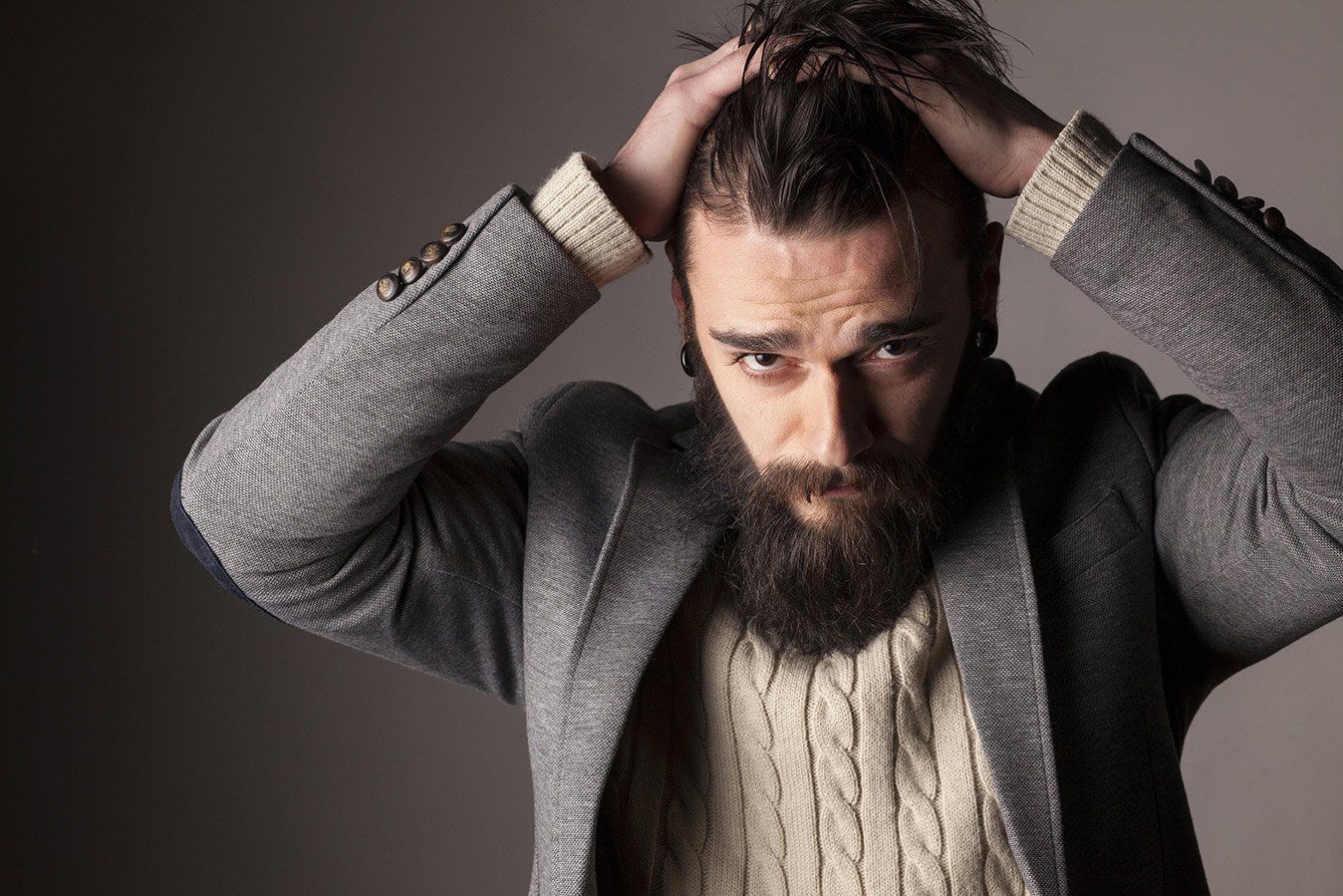Man with a long beard, dark hair pulled back, wearing a gray blazer and cream sweater, hands on head, looking stressed.