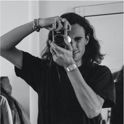Man with long hair, taking a photo with a camera in front of a mirror. He wears a dark shirt and several rings and a watch.