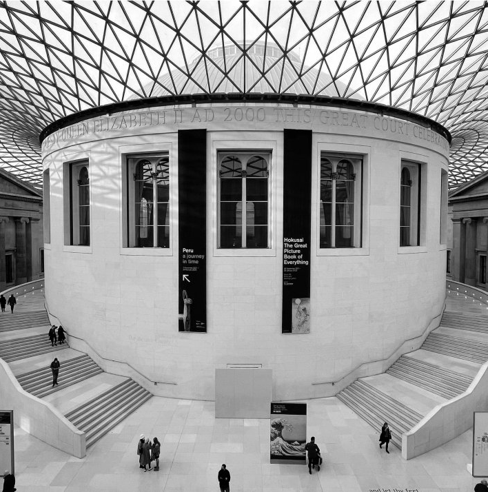 British Museum's Great Court, with people walking. Modern dome roof over a rotunda with windows and stairs.