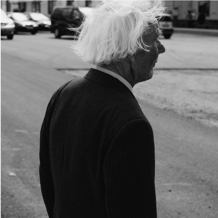 An elderly person with white hair stands on a street, vehicles in the background.