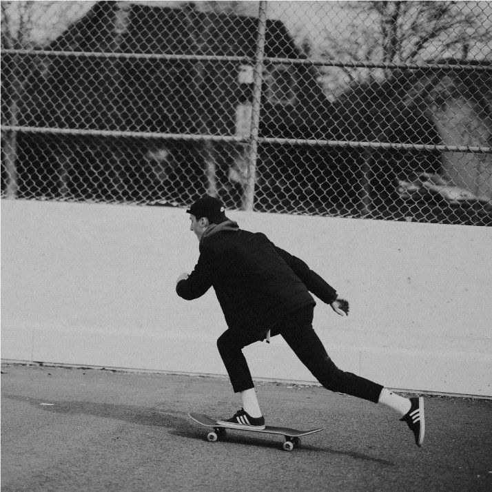 Person in black jacket and cap skateboarding on a paved surface, chain link fence in background.