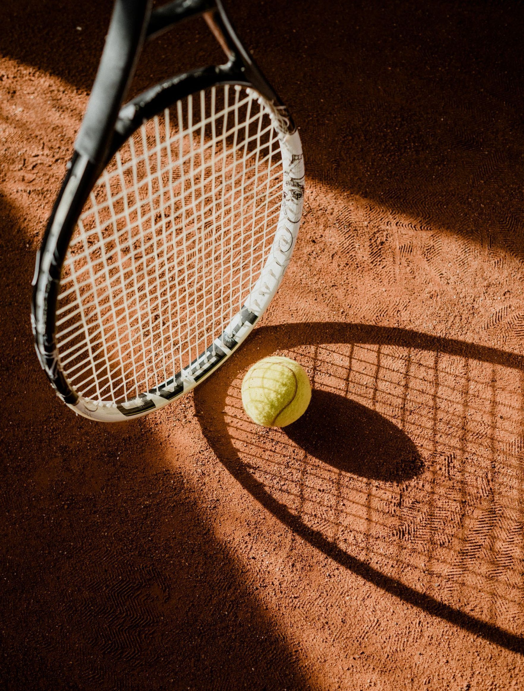 Tennis racket and ball on a clay court, with the racket’s shadow stretching across the surface.