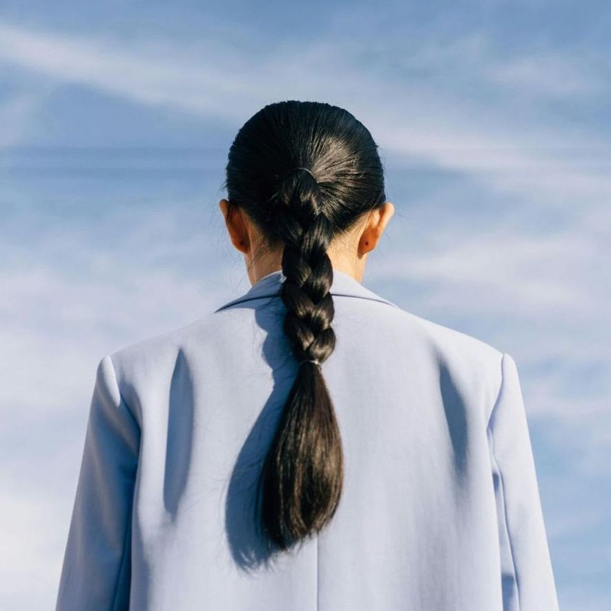 Woman in blue jacket, back to camera, with braided black hair against a blue sky.