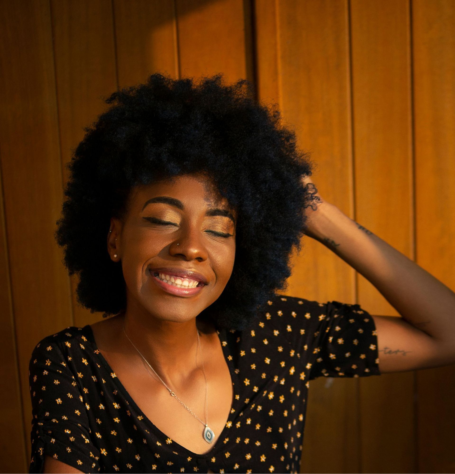 Woman with dark, fluffy hair, smiling with eyes closed, hand in hair, wearing polka-dot top.