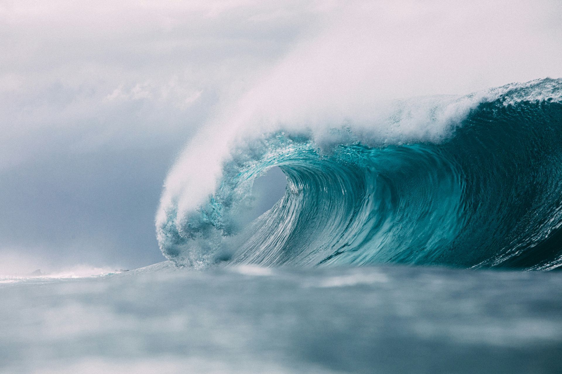 A large, crashing turquoise wave with a foamy crest. Ocean under a cloudy sky.
