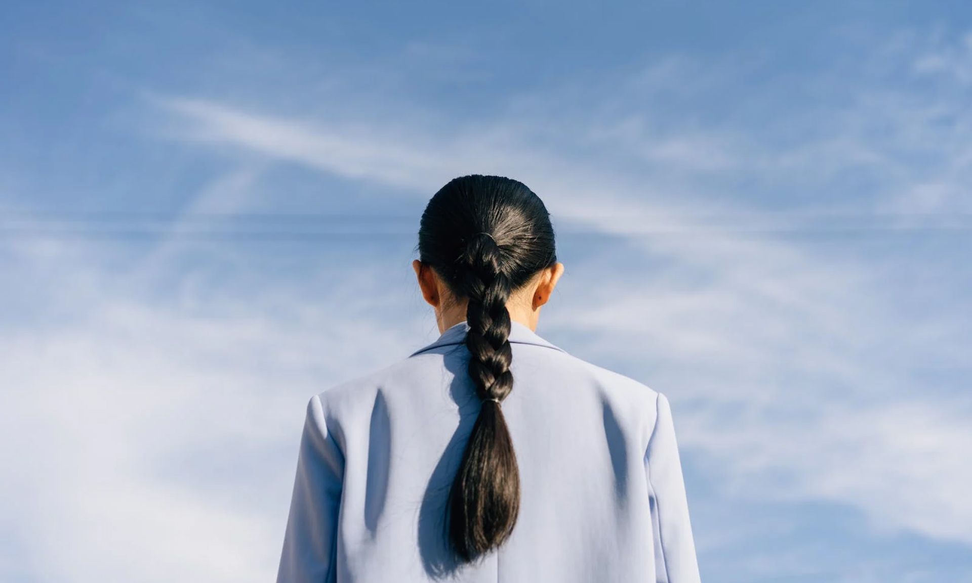 Person with dark hair in a braid, wearing a blue blazer, looking at a bright blue sky with white clouds.