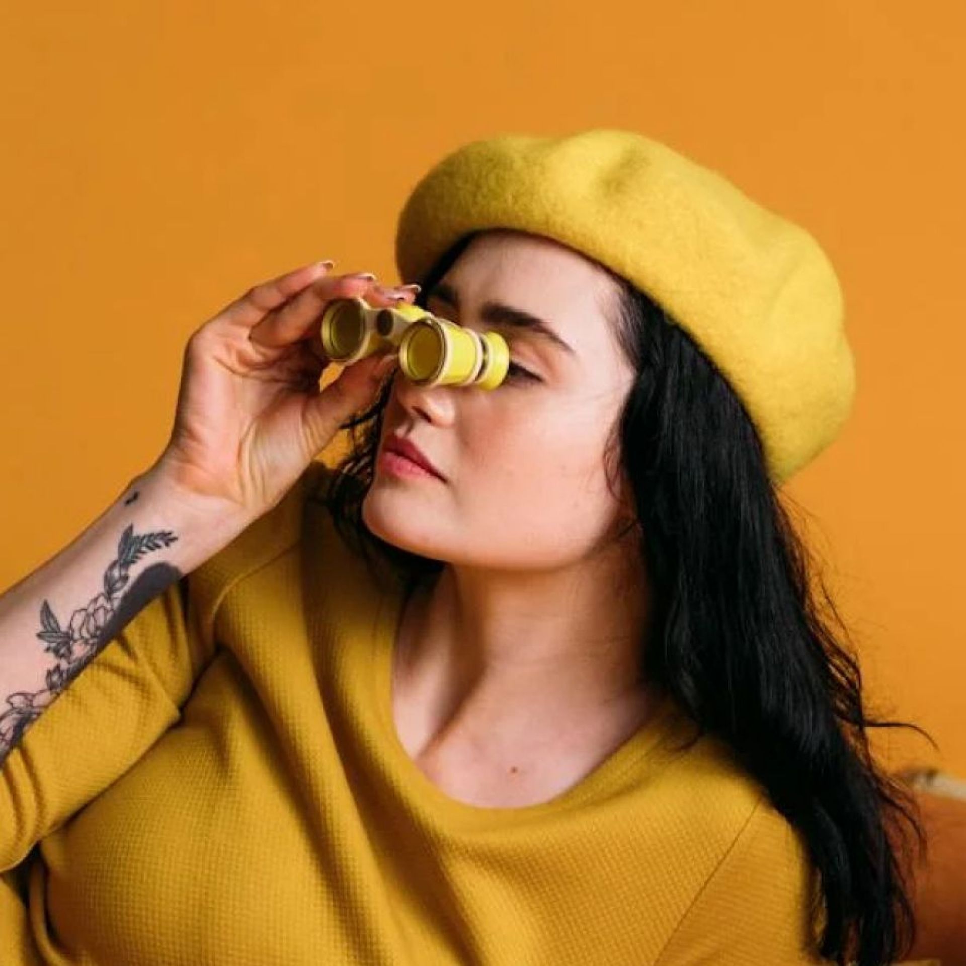 Woman in yellow beret and shirt looking through gold binoculars against an orange background.