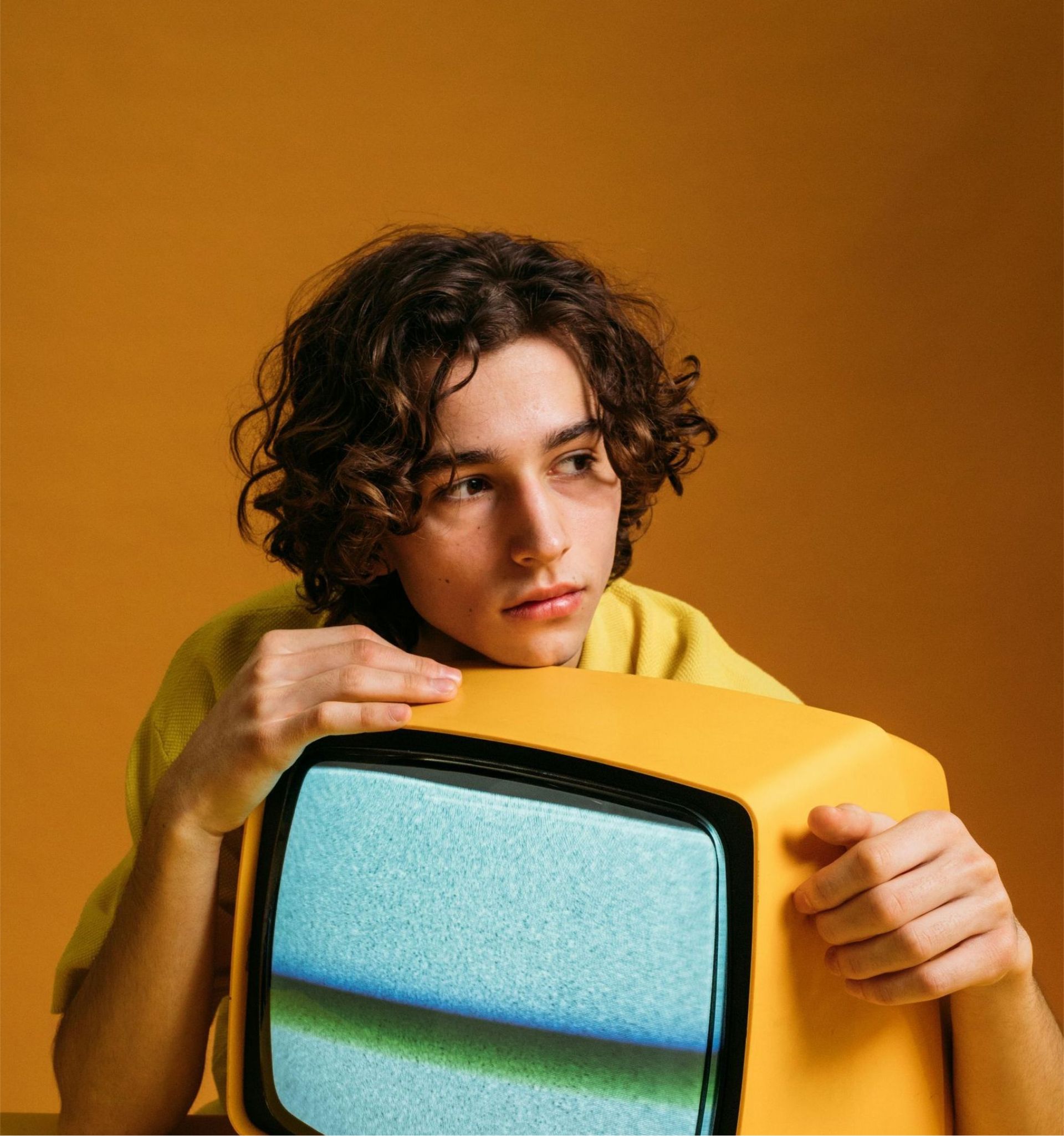 Young person with curly brown hair, resting chin on a yellow vintage television, gazing thoughtfully.